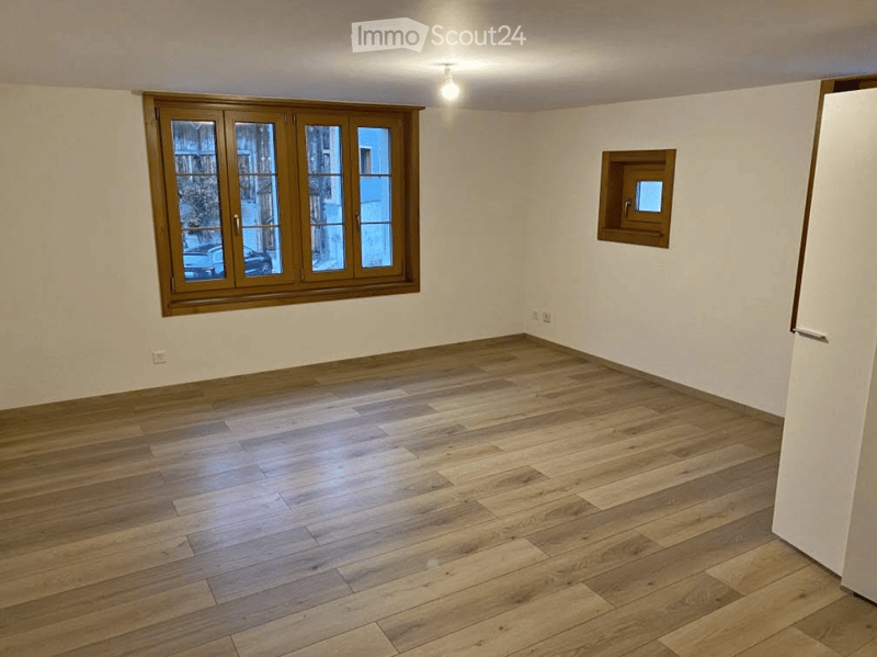Empty room with wooden flooring, large window with wooden frame, small window, and white walls.