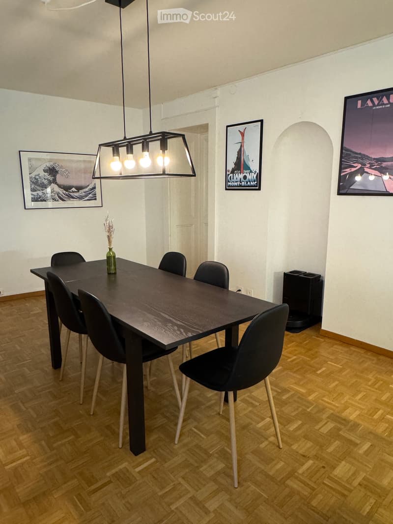 Dining room with a dark wooden table surrounded by six black chairs, a vase with dried flowers, and a modern pendant light hanging from the ceiling. The room features wooden flooring and several framed artworks on the walls.