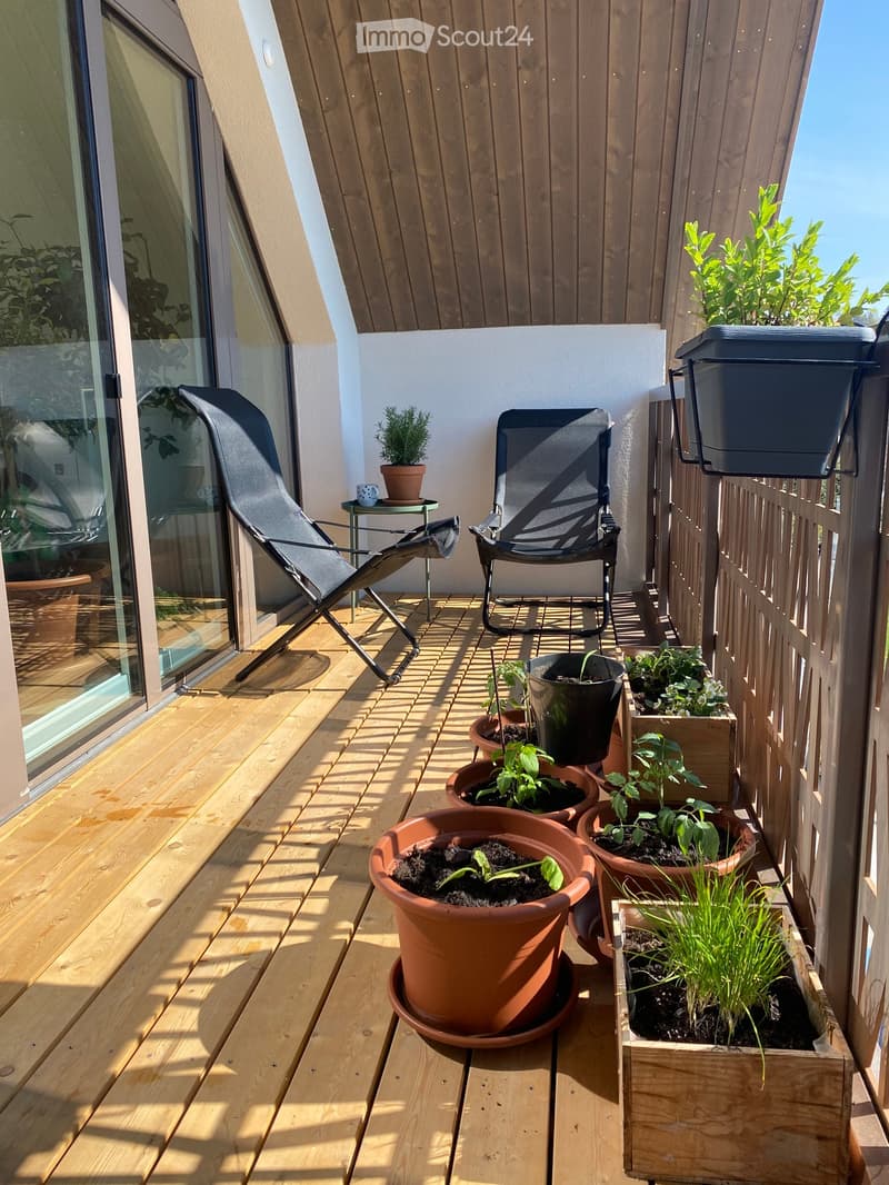 Wooden balcony with potted plants, a small table with a cup, and two chairs, one of which is a lounge chair.