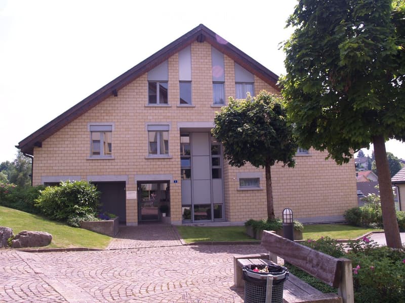 Two-story house with beige brick facade, multiple windows, and a central entrance. The house is surrounded by a well-maintained garden with a paved pathway, a bench, and a trash bin.
