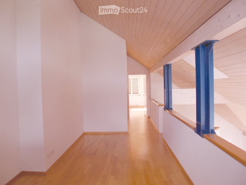 Empty hallway with light wooden flooring, white walls, and a slanted wooden ceiling. Blue structural supports are visible on the right side.