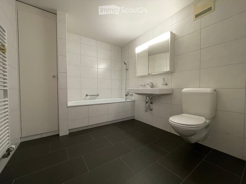 Modern bathroom with white tiles, dark floor tiles, bathtub, toilet, sink, and illuminated mirror.