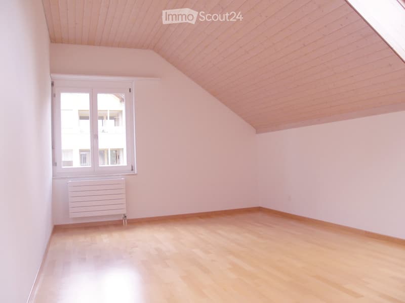 Empty room with light pink walls, wooden floor, and a window with white frame. The ceiling is slanted with wooden paneling.