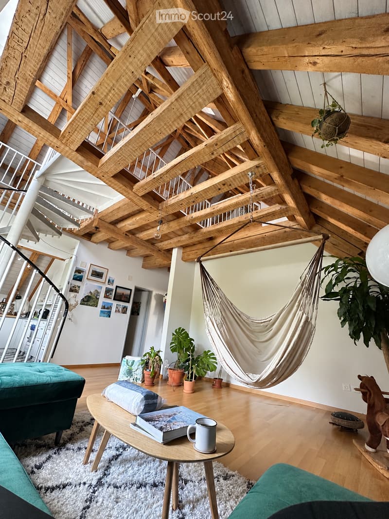A modern living room with exposed wooden beams, a hanging hamaca, a teal sofa, a round coffee table with books and a mug, potted plants, and picture frames on the wall.