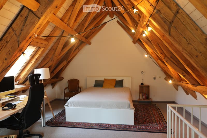 Cozy attic bedroom with wooden slanted ceilings, a white bed with pillows, a desk with a computer, a chair, and a small side table with books.