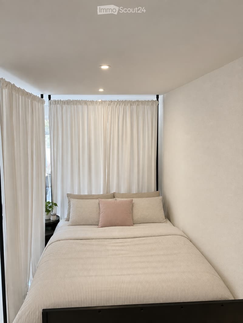 A modern bedroom with a neatly made bed, multiple pillows, a nightstand with a plant, and large windows covered by white curtains.