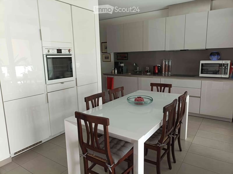 Modern kitchen with white cabinets, stainless steel appliances, a glass dining table with six wooden chairs, and a decorative bowl on the table.