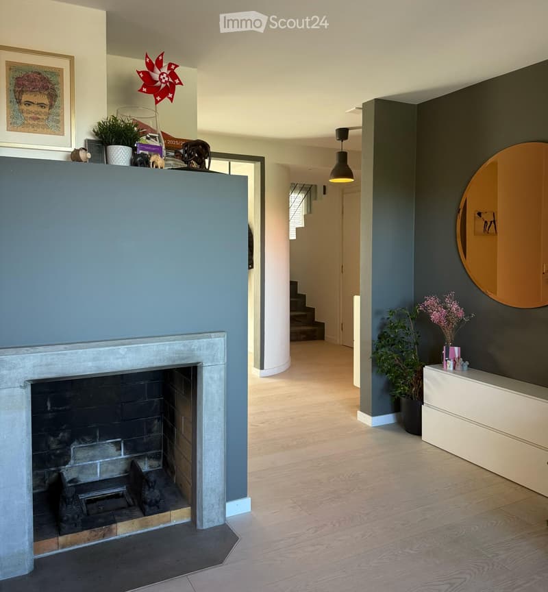 Modern hallway with a fireplace, wooden flooring, and a staircase leading upwards. Features include a round mirror, potted plants, and decorative items on a console table.