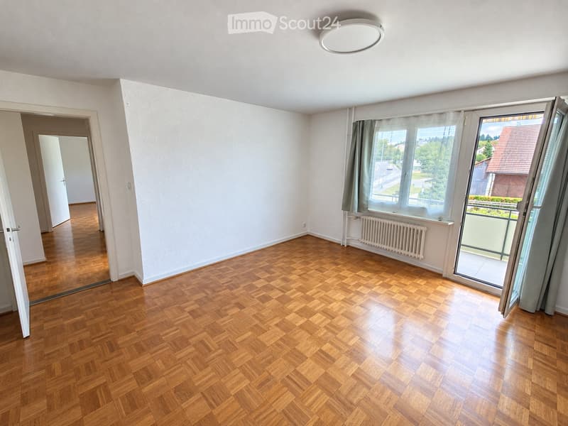 Empty room with wooden herringbone flooring, white walls, large windows with curtains, and a sliding door leading to a balcony.