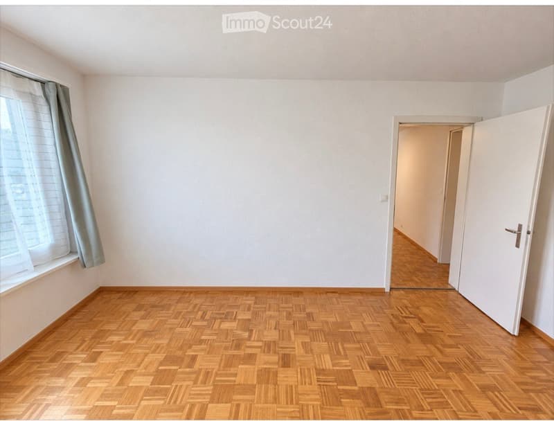 Empty room with wooden parquet flooring, white walls, and a window with a curtain. A doorway leads to another room.