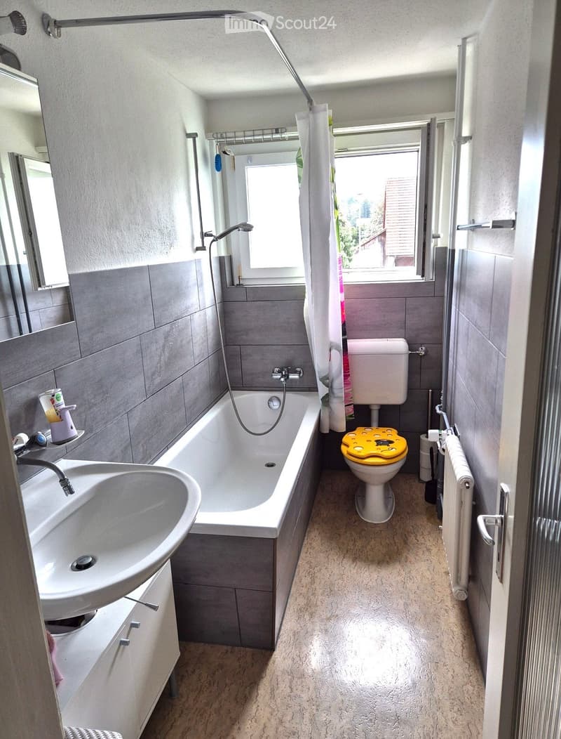 Modern bathroom with a white sink, bathtub, toilet with a yellow seat cover, and a shower area with a curtain. The room features grey tiled walls, a window, and a radiator.