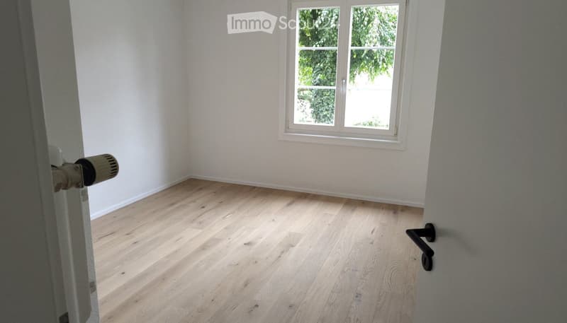 Empty room with light wooden flooring and white walls, featuring a single window with white frame and a door with a silver handle visible on the left side.