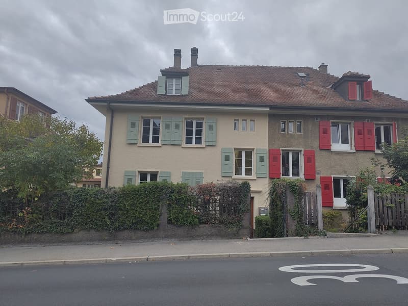 Two-story house with beige facade, multiple windows with shutters, and a well-maintained hedge in front.