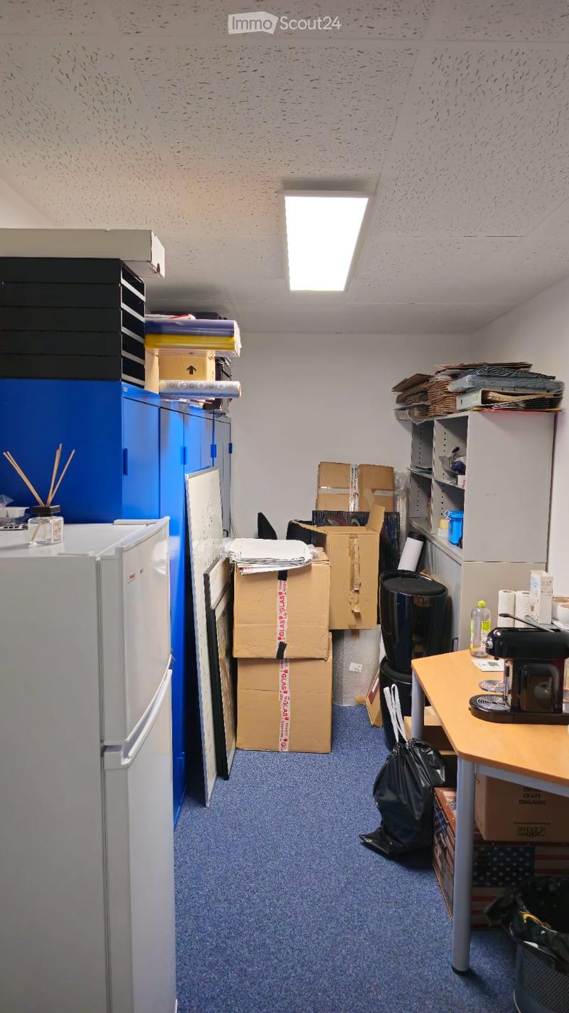 A room with blue carpet, various boxes, storage units, a desk, a printer, a refrigerator, and a trash can. The room appears to be a storage area or office space.