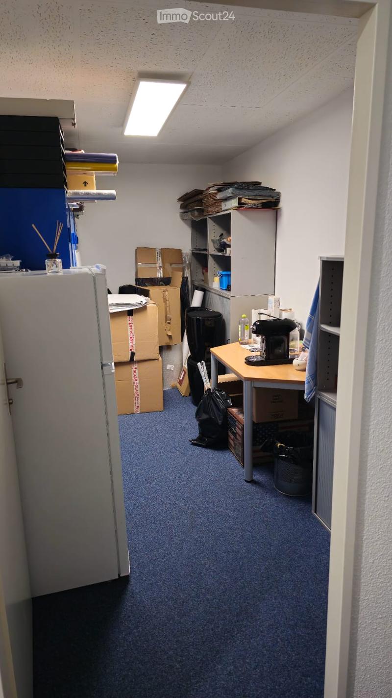 A small, furnished room with a refrigerator, a small table with a coffee maker, shelves holding various items, and several cardboard boxes stacked around the space.