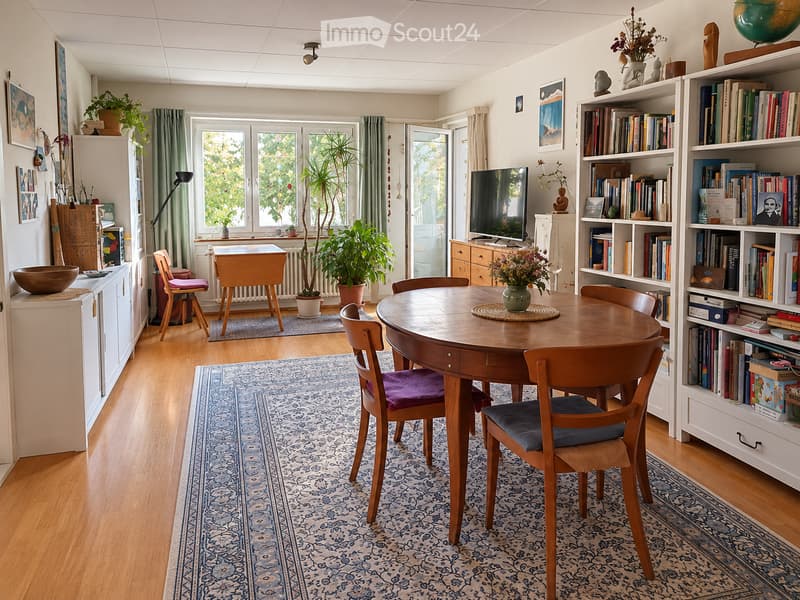 Brightly lit room with wooden flooring, a round wooden dining table surrounded by chairs, a bookshelf filled with books, a television, potted plants, and decorative items.