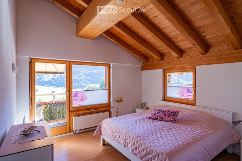 Cozy bedroom with wooden ceiling beams, large windows offering a scenic mountain view, a neatly made bed with pink bedding, a nightstand with a lamp, and a small table with decorative items.