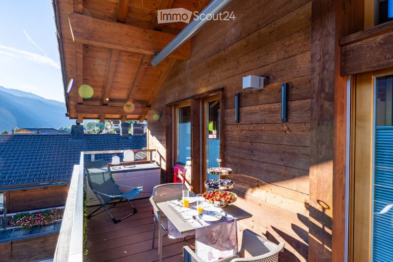 Wooden balcony with a dining setup, overlooking a scenic mountain view. Includes a table with chairs, cups, and dishes.