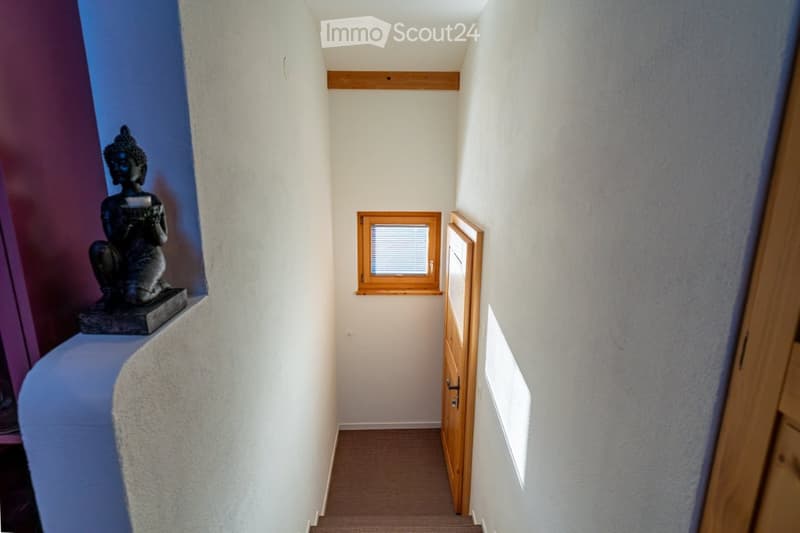 Narrow hallway with light-colored walls, wooden door, small window, and a decorative statue on a shelf.