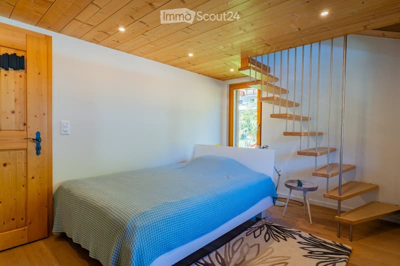 Cozy bedroom with wooden ceiling, bed with blue blanket, small stool, and a wooden staircase leading upwards.