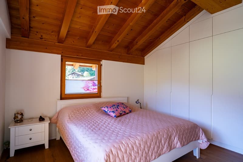 Cozy bedroom with wooden ceiling beams, white walls, a large window, a bed with pink bedding, a nightstand, and built-in white wardrobes.