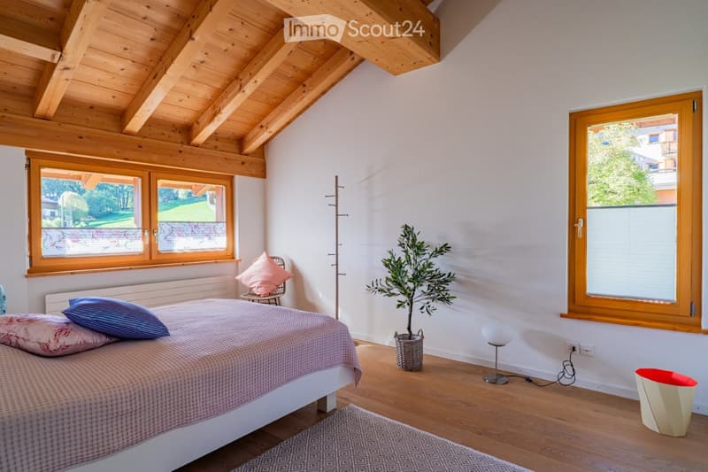 Cozy bedroom with wooden ceiling beams, large windows, a bed with pillows and a blanket, a chair with a cushion, a potted plant, and a floor lamp.