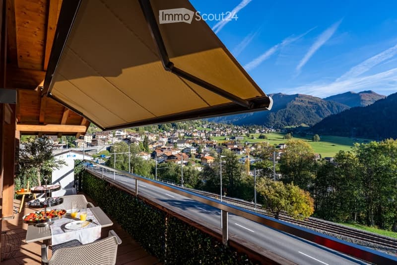 Balcony with a scenic view of a town and mountains, featuring a small table with chairs, a bowl of fruit, and a cup.