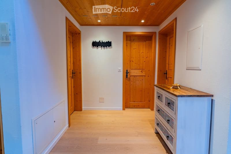 A clean, well-lit hallway with wooden flooring, white walls, and several wooden doors leading to different rooms. A small console table with drawers is positioned on the right side, and a modern light fixture is mounted on the wall.