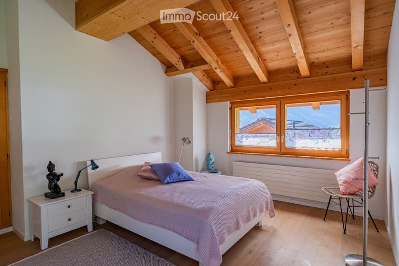 Cozy bedroom with wooden ceiling beams, large window, bed with pink and blue bedding, nightstand with lamp, decorative items, and a wicker chair.