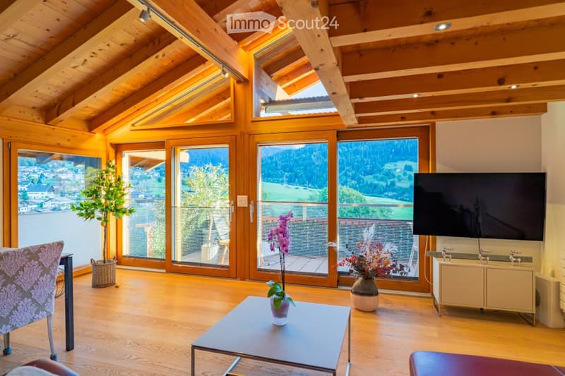 Bright living room with wooden beams, large windows, and a scenic mountain view. Features a sofa, coffee table, television, and decorative vases.