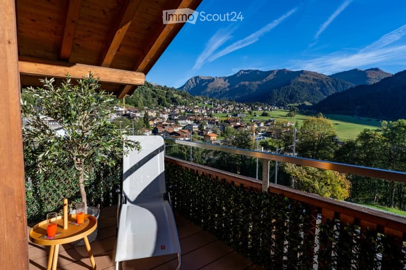 Balcony with wooden decking, small round table with drinks, white lounge chair, potted plants, and scenic mountain view of a town.