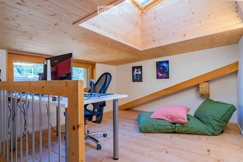 A cozy attic room with wooden beams and skylights, featuring a desk with a computer, a comfortable chair, and a small sofa with pillows.