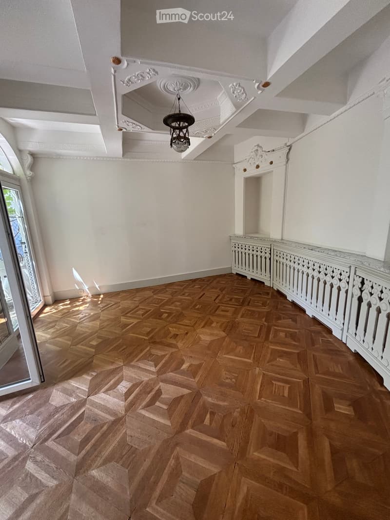 Elegant hallway with ornate white walls, a decorative chandelier, and a polished wooden floor with geometric patterns. Features a white balustrade and a large window allowing natural light to enter.