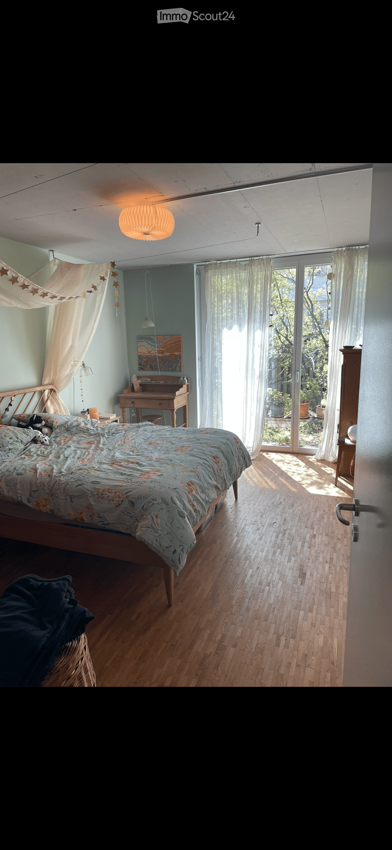 Bright bedroom with wooden flooring, large window with sheer curtains, bed with floral duvet, nightstand, desk, and artwork on the wall.