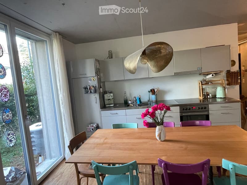 Modern kitchen with light grey cabinets, stainless steel appliances, a wooden dining table with colorful chairs, and a vase of pink flowers.