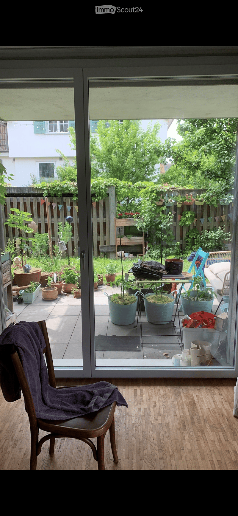A wooden chair with a purple blanket draped over it is positioned near a large sliding glass door that opens to a garden area filled with potted plants and outdoor furniture.
