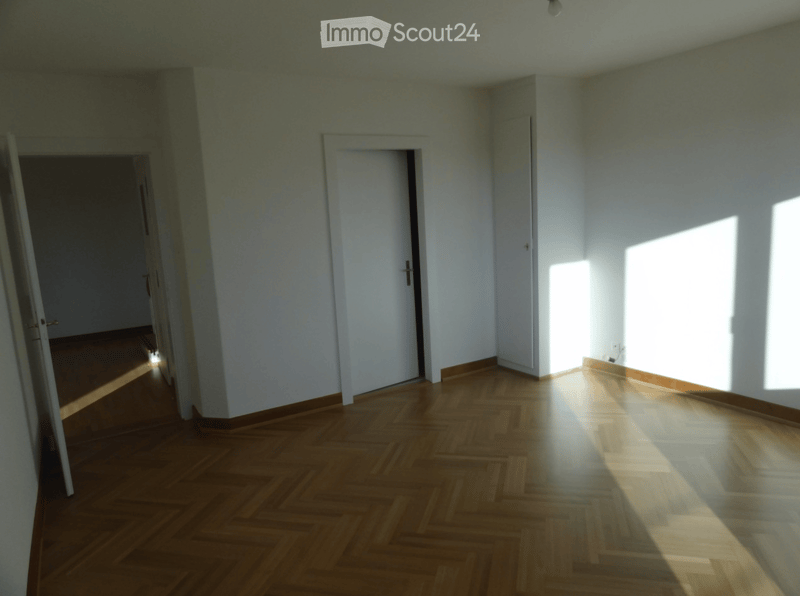 Empty room with wooden parquet flooring, white walls, and natural light coming through a window on the right side. Two doors are visible, one leading to another room on the left and another closed door in the center.