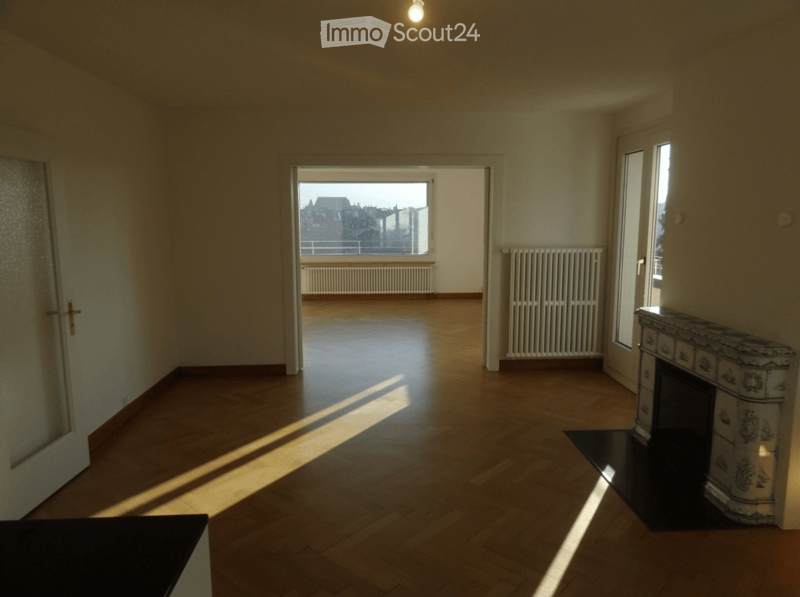Empty room with wooden flooring, large windows, and a decorative fireplace. Natural light streams in from the windows.