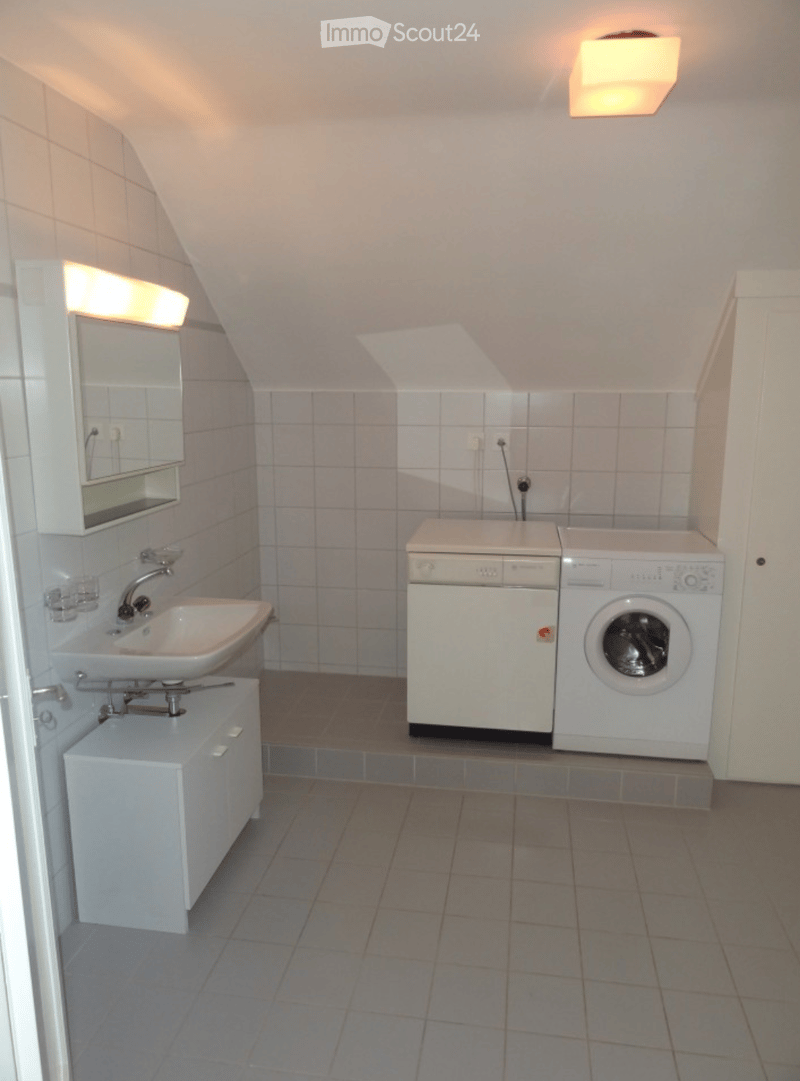Bathroom with a sink, mirror, dishwasher, washing machine, and a small cabinet. The room has tiled walls and a slanted ceiling typical of an attic space.