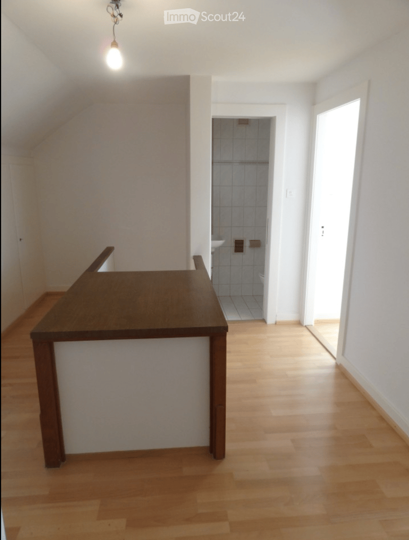 Empty room with wooden flooring, white walls, and a small table in the foreground. A bathroom is visible in the background.