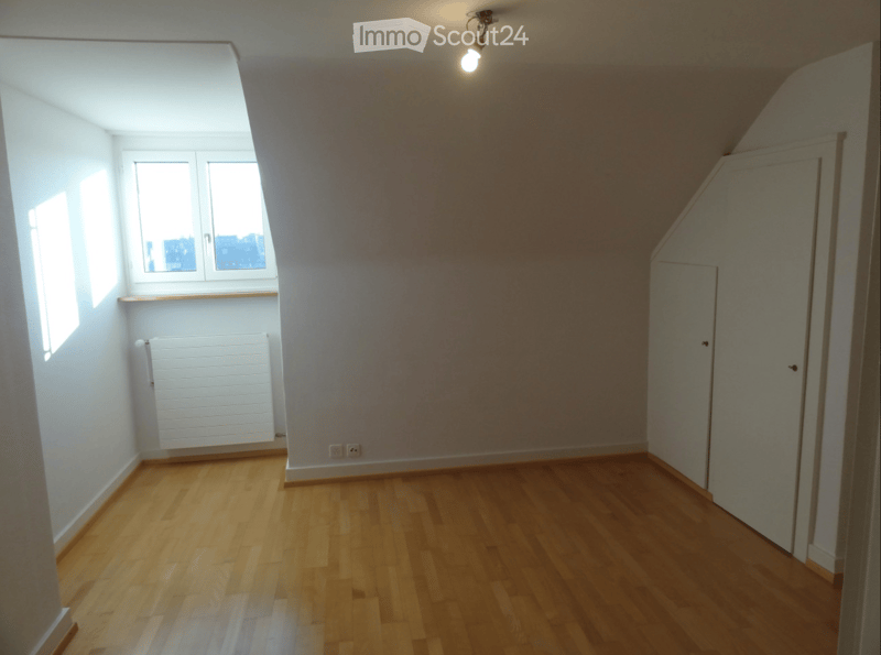 Empty attic room with wooden flooring, white walls, and a window with a radiator below it.