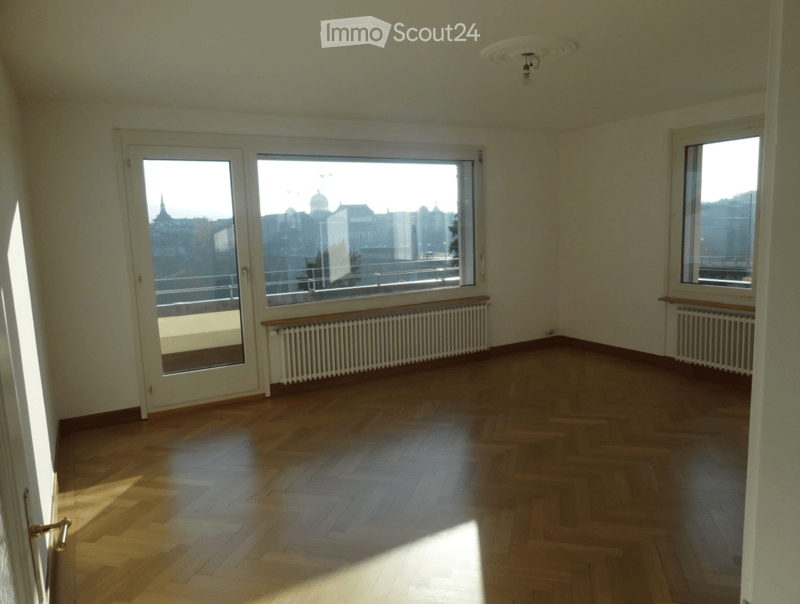 Empty room with wooden flooring, large windows offering a cityscape view, and radiators along the walls.