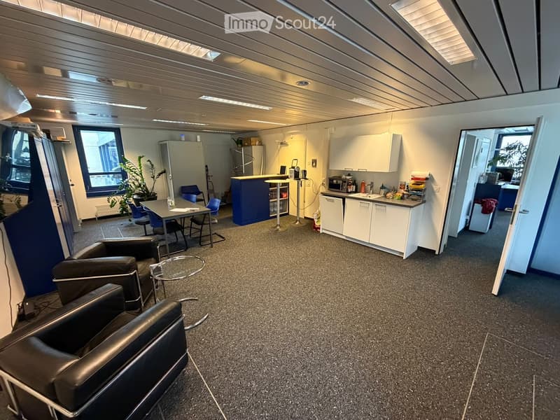 Modern office space with a seating area, kitchen area, and work tables. Features include a black leather sofa, round glass table, blue chairs, and a small kitchen with cabinets and appliances.