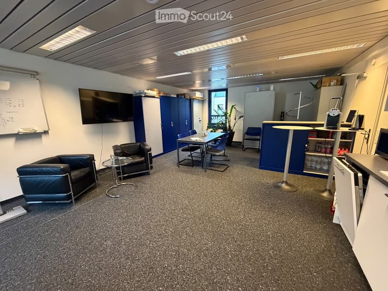 Modern office or meeting room with a sofa, chairs, a table, a screen, a small bar area, and a plant. The room has a carpeted floor and white walls.