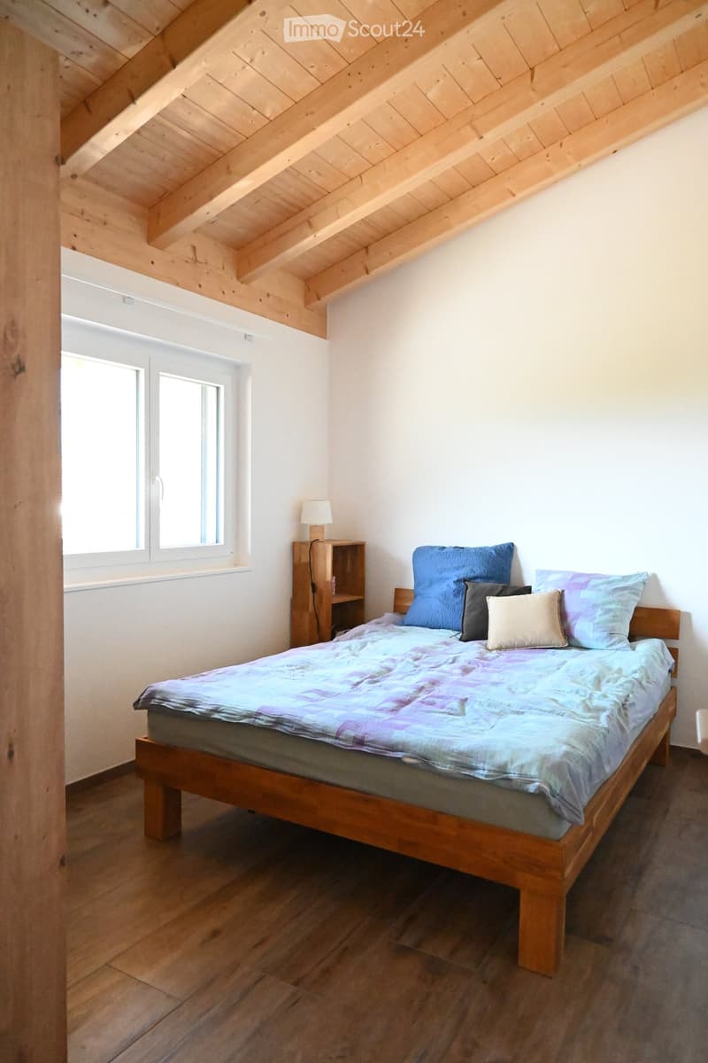 Cozy attic bedroom with wooden slanted ceilings, a wooden bed with colorful bedding, a nightstand with a lamp, and a small shelf.