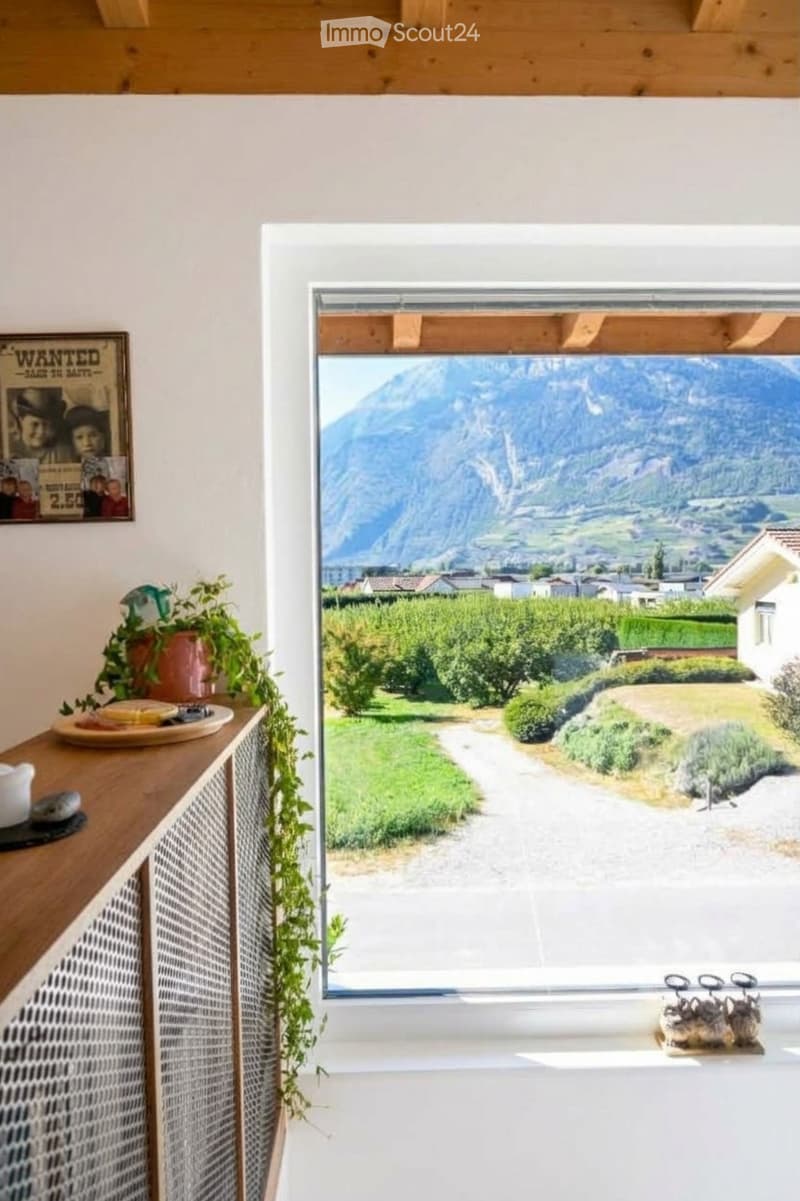 Bright room with large window offering a scenic mountain view, featuring a wooden cabinet with a plant, a plate, and a teapot on top.