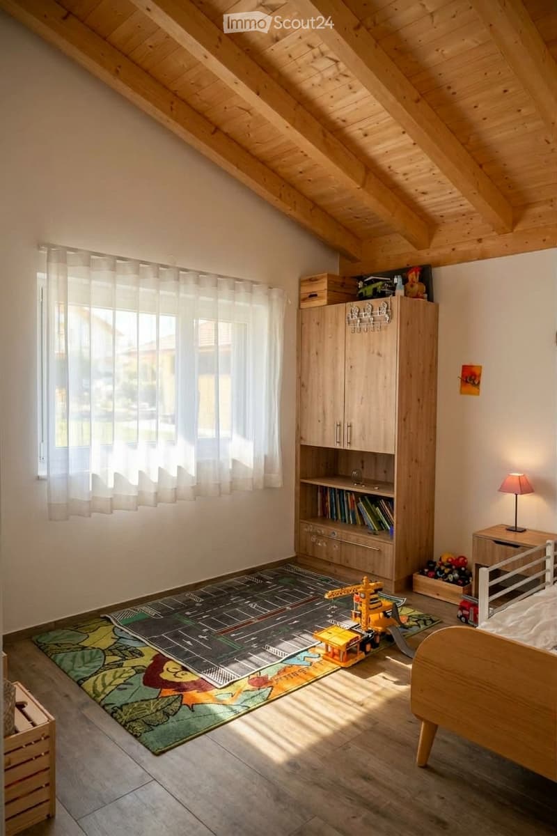 Bright, wooden-floored bedroom with slanted wooden ceiling, large window with sheer curtains, bed, wardrobe, bookshelf, table lamp, and children's toys on the floor.