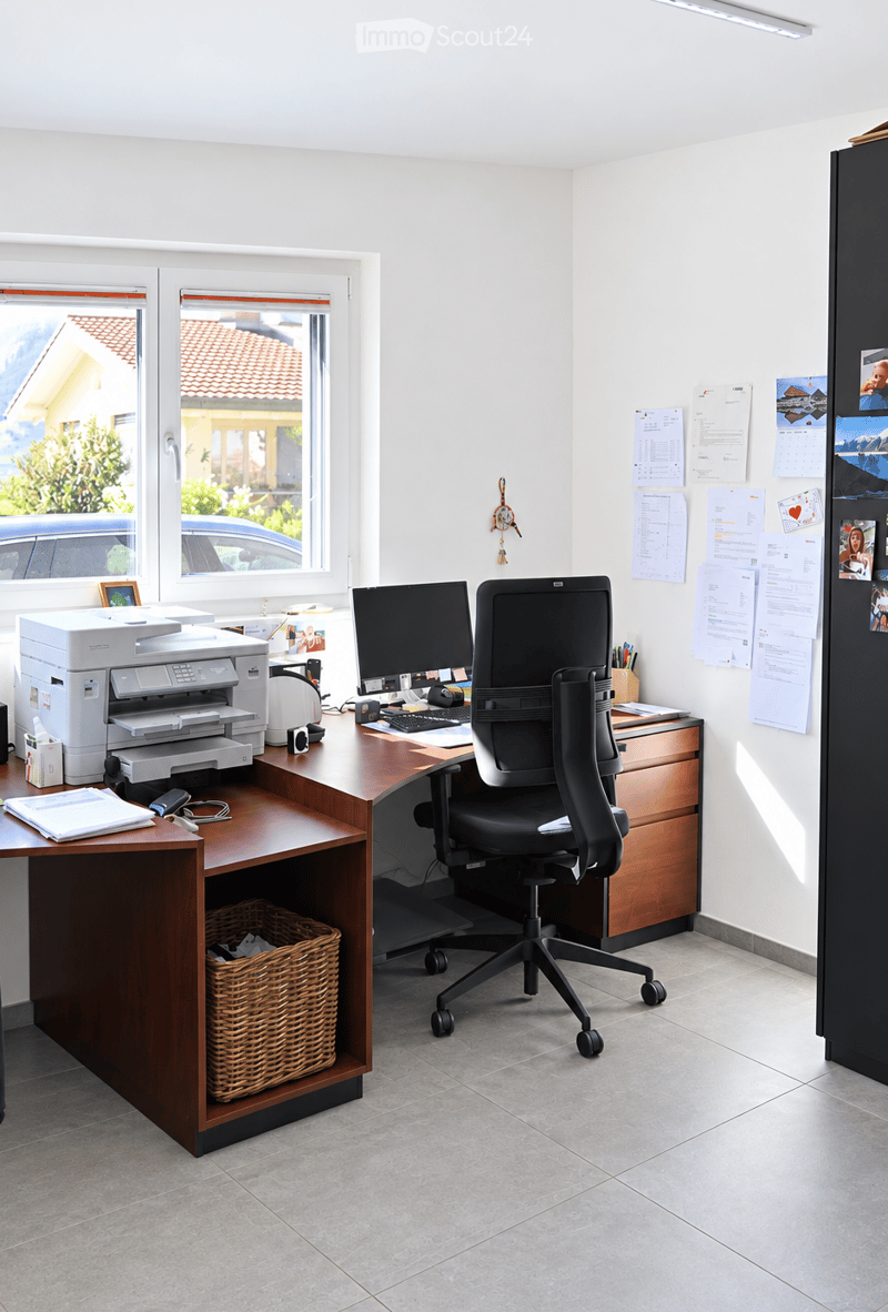 A well-organized office space with a wooden desk, ergonomic chair, computer setup, printer, and various office supplies. The room features large windows with a view of a neighboring house and a parked car.