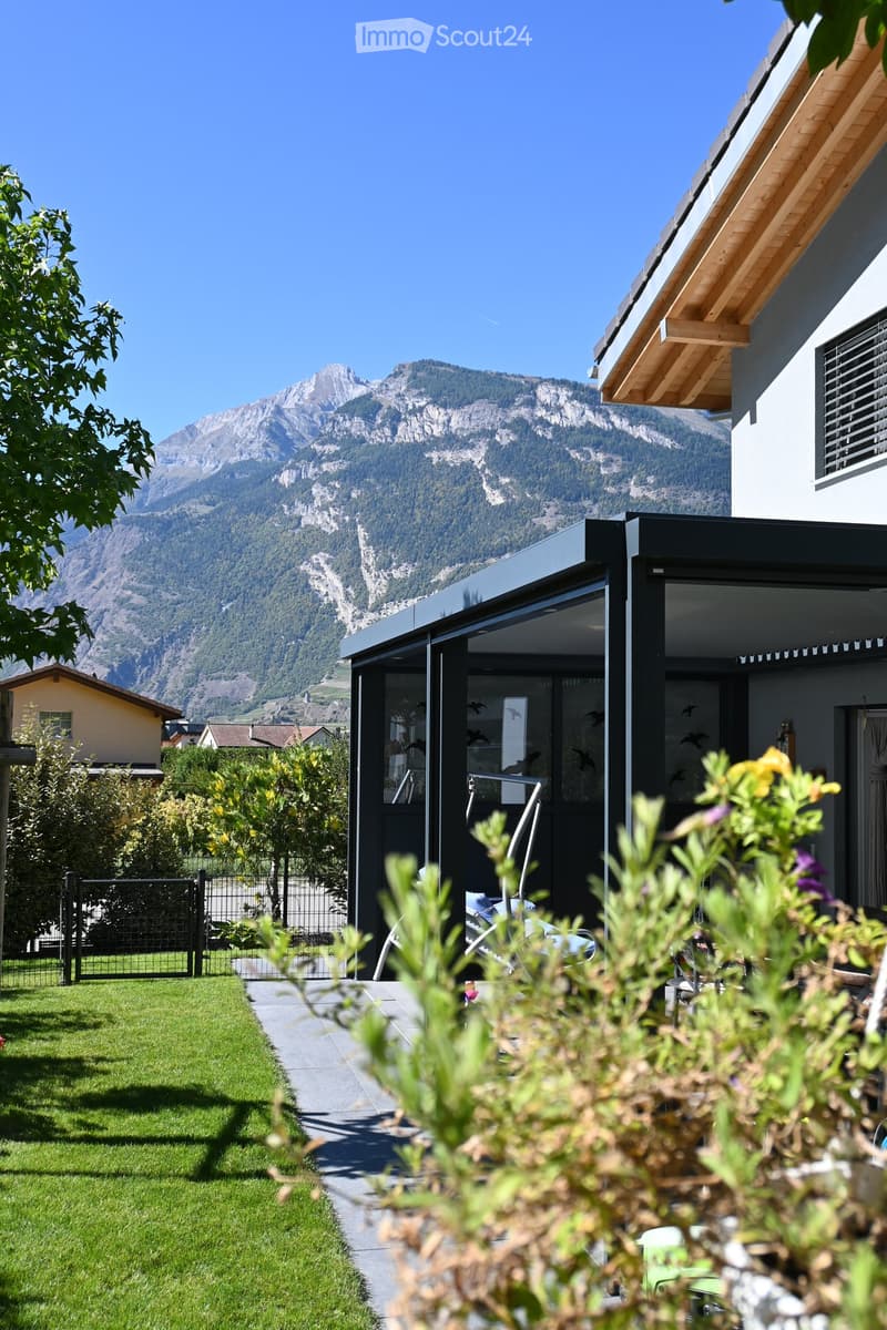 Garden area with a well-maintained lawn, a paved pathway, and a modern outdoor structure with large glass panels. The background features a stunning mountain view.