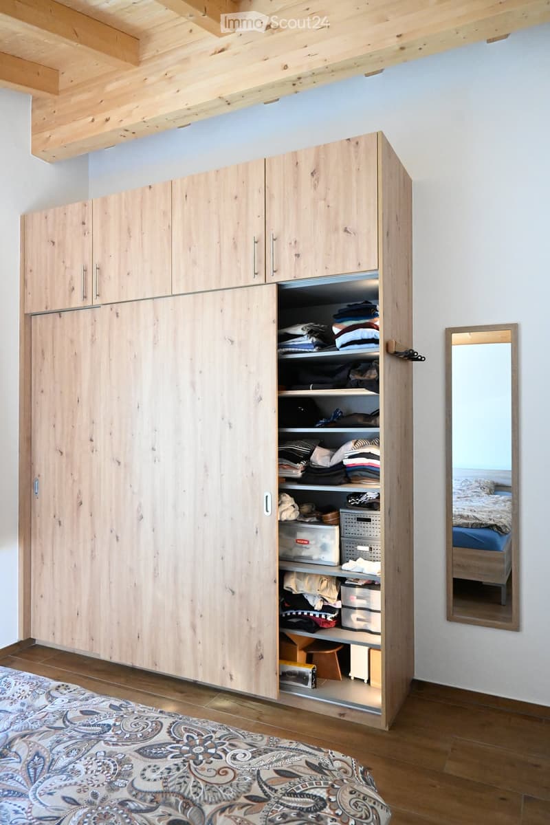A modern bedroom with a large wooden wardrobe, a mirror, a bed with patterned bedding, and a decorative rug on the floor.